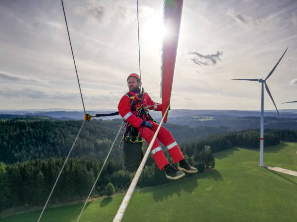 Epic view on charismatic Industrial climber,  rope access technician working on tip of the wind turbine blade with sunset and windfarm behind him, looking away stock photo