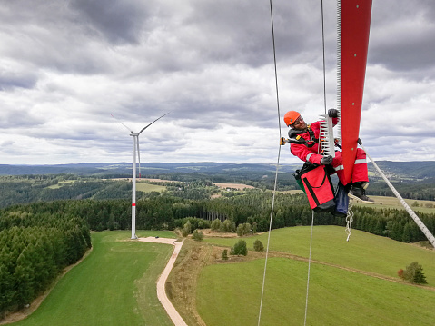 Epic view on Industrial climber,  rope access technician working on tip of the wind turbine blade fasten serrations on trailing edge and with windfarm is behind him, dramatic sky