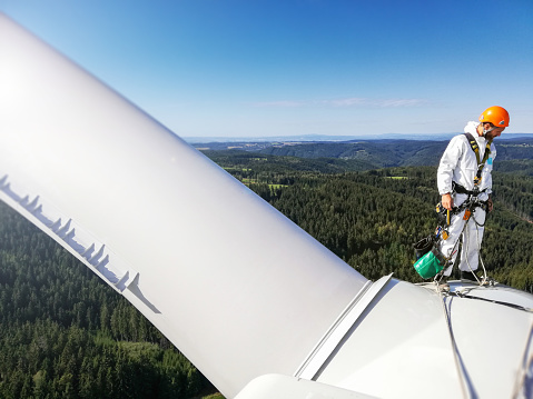 Professional rope access technician - industrial climber standing on roof (hub) of wind turbine and looking down. View on forest behindhim.