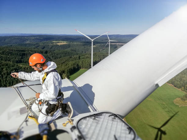 Professional rope access technician kneeling on roof (hub) of wind turbine and is tied up with rope before drop on blade. Epic view on wind-farm behind him. stock photo