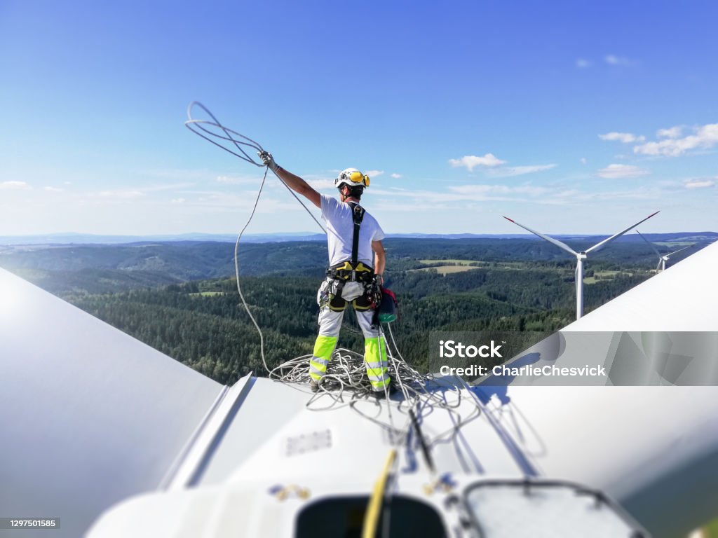 Rear view on Professional rope access technician standing on roof (hub) of wind turbine and pulling rope up. Sun is behind wind turbine. Professional rope access technician standing on roof (hub) of wind turbine and pulling rope up. Sun is behind wind turbine. Wind Turbine Stock Photo