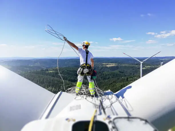 Professional rope access technician standing on roof (hub) of wind turbine and pulling rope up. Sun is behind wind turbine.