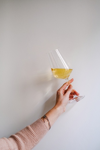 A female hand is holding a glass of white wine on the plain white wall, but the background is full with light and shadow, the wine and the glass give sun beams and reflections