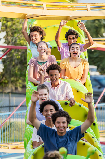 A multi-ethnic group of eight teenagers having fun riding a rollercoaster at an amusement park, smiling and laughing with their arms raised.
