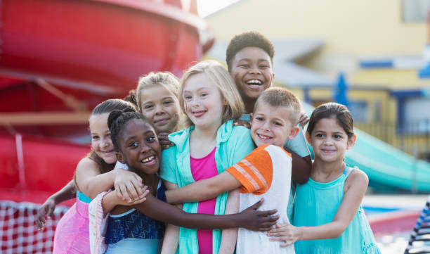 Girl with down syndrome, friends at water park hugging A 10 year old girl with down syndrome having fun at a water park, in the center of a multi-ethnic group of friends, getting a group hug. They are smiling at the camera. male swimsuit standing arm around stock pictures, royalty-free photos & images