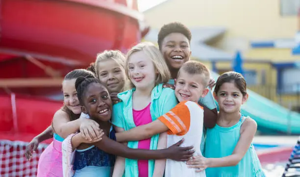 A 10 year old girl with down syndrome having fun at a water park, in the center of a multi-ethnic group of friends, getting a group hug. They are smiling at the camera.