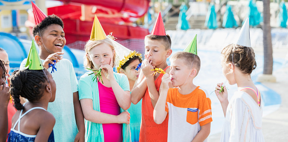 A multi-ethnic group of 8 to 11 years old, having fun together, at a birthday party at a water park. They are wearing party hats, laughing, with their party horns. The girl wearing a pink swimsuit has down syndrome.