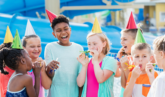 A multi-ethnic group of 8 to 11 years old, having fun together, at a birthday party at a water park. They are wearing party hats, laughing, with their party horns. The girl in the middle wearing a pink swimsuit has down syndrome.