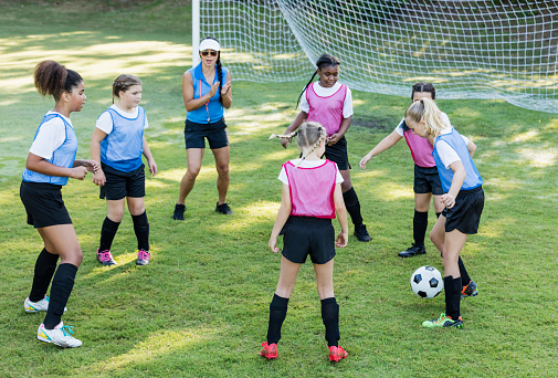 A girl's soccer team on the field in front of the goal, practicing with their coach. The multi-ethnic group of six players, 9 and 10 years old, are standing in a circle, passing the soccer ball to one another.