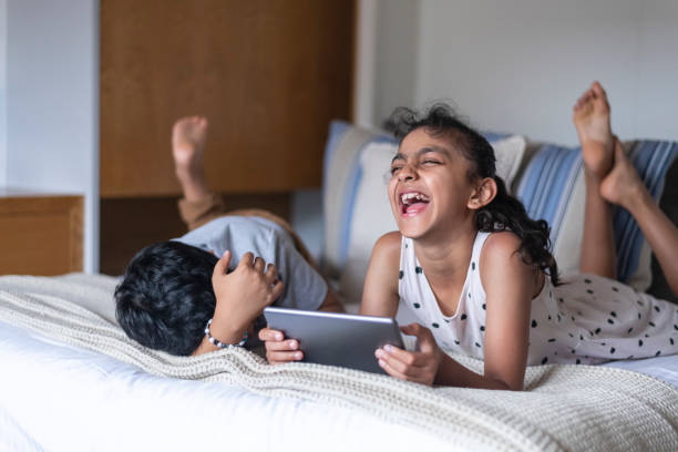 Affectionate elementary age siblings laughing while doing screen time on tablet computer Happy elementary age brother and sister laugh while watching a video or playing a game on a tablet computer at home. Screen time, gaming, and family lifestyle concepts. facebook news australia stock pictures, royalty-free photos & images