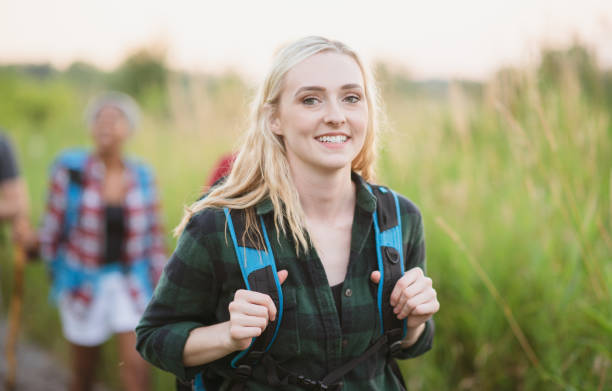 A young female adult of Caucasian ethnicity is outdoors hiking on trails with some friends. She is carrying a backpack and is smiling at the camera.