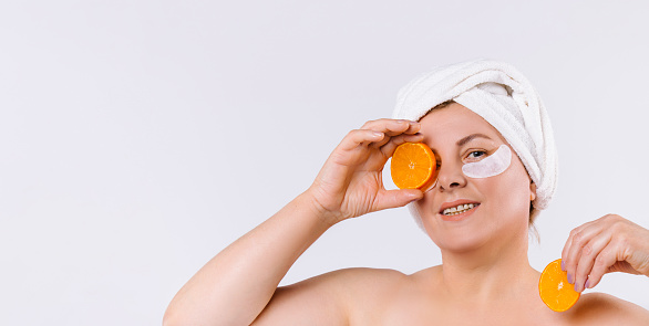 Banner- long format. Beautiful smiling elderly woman with eye patches, looking to camera, holding fresh orange slice. Skinimalism care, spa, natural beauty and cosmetology concept. High quality photo