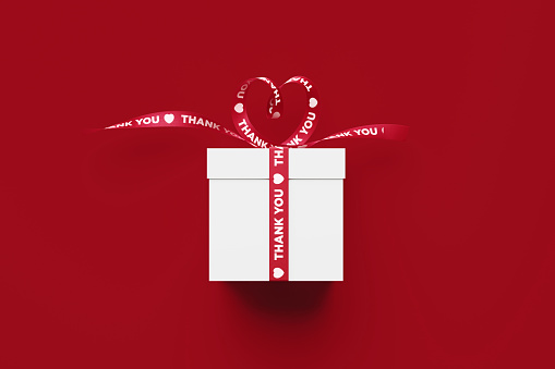 White gift box tied by heart shaped and thank you printed red ribbon sitting over red background. Horizontal composition with copy space. Directly above. Great use for Christmas, Mother's Day, birthday and Valentine's Day related gift concepts.