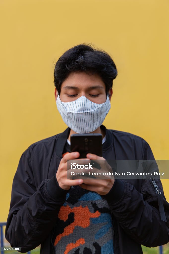 A Young Man With Mask Using His Mobile Phone Young latin man with mask is using his phone. Yellow Background Adult Stock Photo