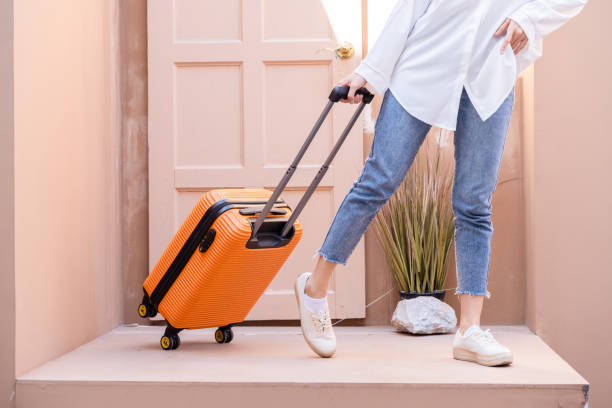 młoda samotna podróżująca kobieta stojąca z gruzińskimi drzwiami, gdy holuje swoją jasnopomarańczową walizkę wypełnioną zdalną technologią pracy, odkładając bilety lotnicze w lewej kieszeni - shoe leaving women summer zdjęcia i obrazy z banku zdjęć
