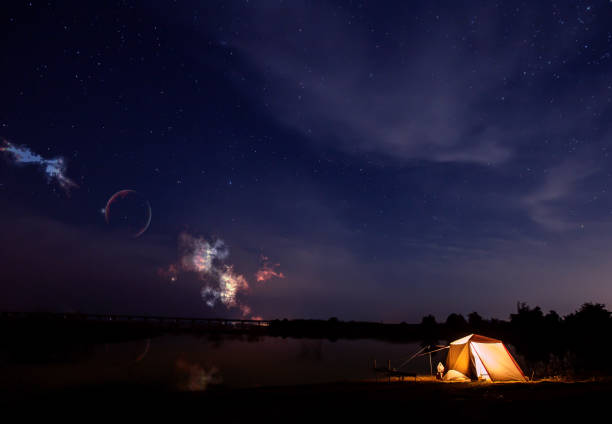 Traveling and camping concept - camp tent at night under a sky full of stars. Orange illuminated tent. Beautiful nature - field, forest, plain. Moon and moonlight
