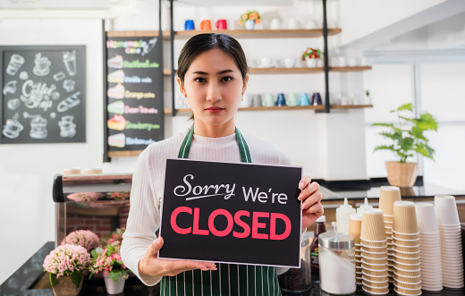 Unhappy barista woman in apron and holding closed shop signboard in cafe. Portrait of sorry asian young girl standing to front counter in coffee shop with show closed signboard because service timeout