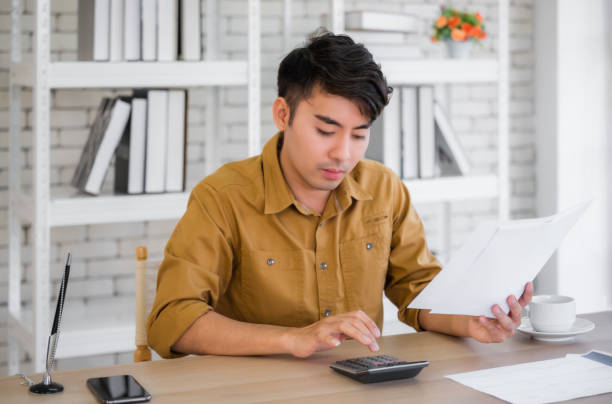 istock Asian businessman calculating checking balance and costs by using calculator in workplace. Young male sitting at desk and doing paperwork for pay taxes. 1297531066
