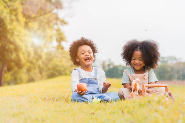 deux enfants africains foncés de peau garçon et fille se reposant et mangeant des fruits dans les parcs et à l’extérieur avec heureux. garçon et fille africains d’enfant mangeant la pomme et la banane avec le sourire dans l’automne tropical de d - smiling little girls little boys autumn photos et images de collection