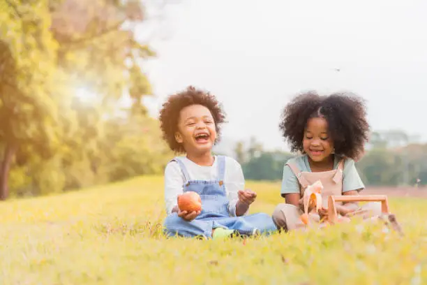 Photo of Two african dark skin child boy and girl sitting and eating fruits in parks and outdoors with happy. African child boy and girl eating apple and banana with smile in grass field tropical autumn.