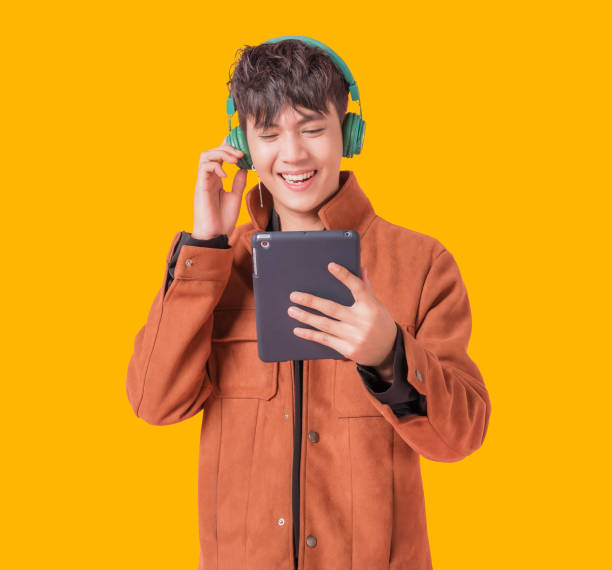 Asian handsome man smiling and listening music by big green headphones by smartphone search Asian handsome man smiling and listening music by big green headphones by smartphone search. Happy boy from listen to music using bluetooth technology in to smartphone isolated on yellow background. asian man headphones stock pictures, royalty-free photos & images