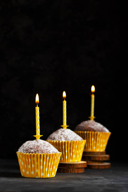 istock Three cupcakes in a paper cup with a yellow candle with a flame on a dark background on wooden coasters birthday card 1297532884