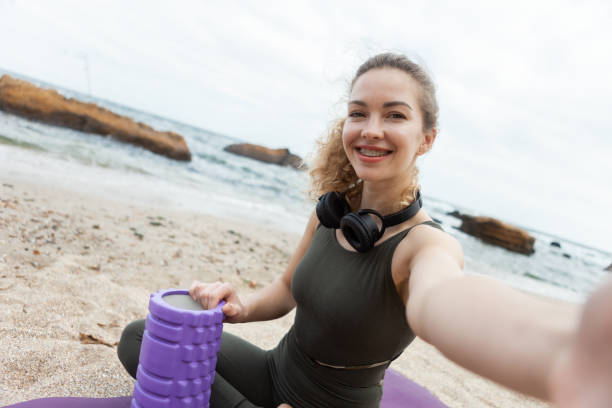 istock Fit woman with massage roller makes selfie while sitting on the mat. Outdoor training. 1297545087