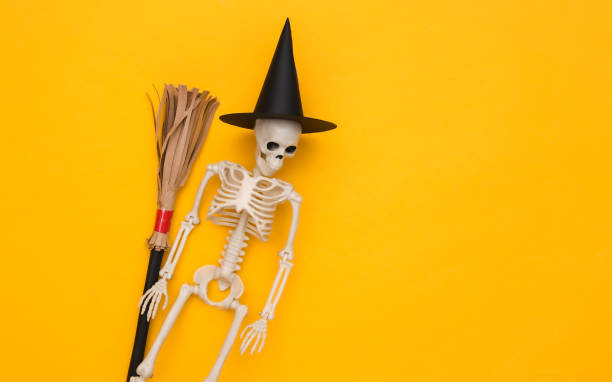 istock Skeleton in a witch's hat and broomstick. Yellow background. Halloween minimalistic concept. Top view. Flat lay 1297551298