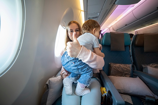 concept everything is OK. little cute toddler jumping on her knees with a young beautiful mother in an airplane chair. mom shows thumb up and looks at the camera with a smile