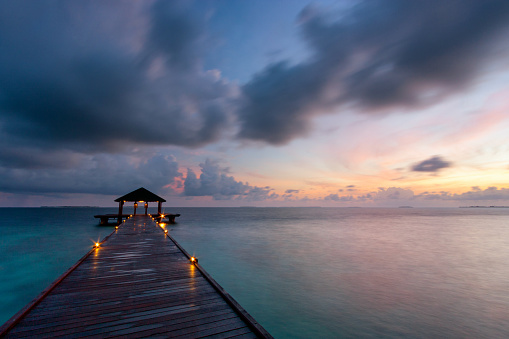 Dawn in the Maldives, and the sky above a wooden pier lights up with the first rays of sunlight.