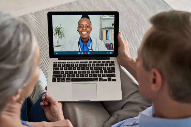 Photo of Over shoulder view of old grandparents couple patients video calling virtual doctor using laptop at home. Online telemedicine chat meeting. Seniors ehealth, telehealth consultation, tele medicine.