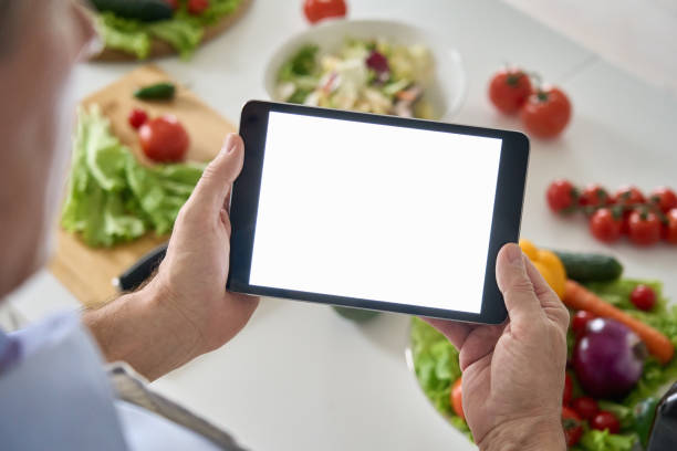 Senior man holding in hands using tablet computer mockup screen on healthy food vegetable background. Online grocery shopping delivery app ads concept, cook book diet plan recipes, over shoulder view.