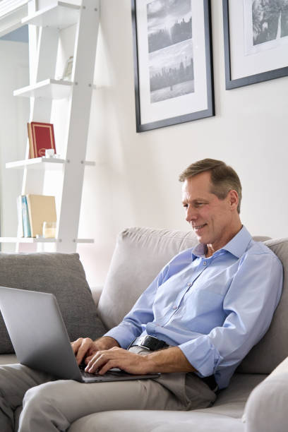 istock Smiling mature 60s older senior man typing on laptop computer tech device sitting on sofa, working or e learning online, using banking applications, buying insurance, ordering food on website at home. 1297569949