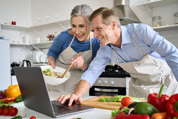 Happy old middle aged 50s couple using laptop computer preparing healthy food diet vegetable salad at home together, searching recipes, ordering shopping online, watching cooking class in kitchen. feliz pareja de mediana edad de los años 50 usando computadora portátil preparando ensalada de verduras de dieta de alimentos saludables en casa juntos, buscando recetas, ordenando compras en línea, viendo la clase de cocina en la cocina.