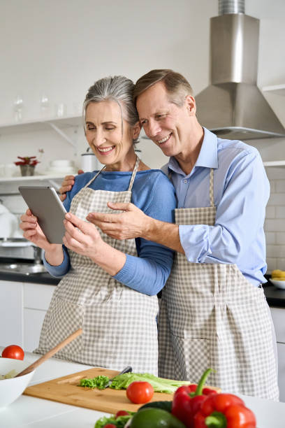 Smiling older senior 60s vegan couple preparing fresh vegetable salad in kitchen using tablet ordering food in grocery e shop or supermarket doing ecommerce shopping online cooking together at home.