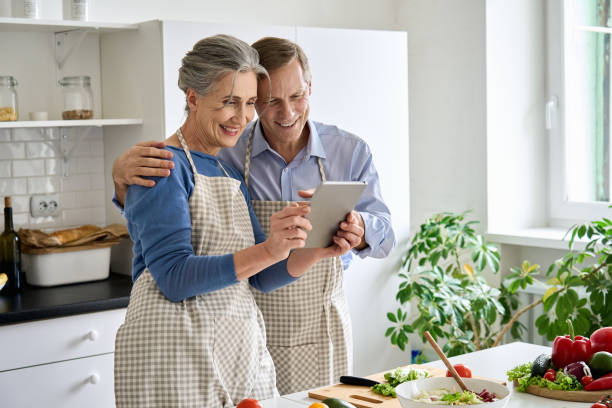 istock Happy middle aged 50s couple preparing salad using digital tablet in kitchen. Smiling mature older husband and wife holding computer cooking together searching healthy diet food online recipes at home 1297569971