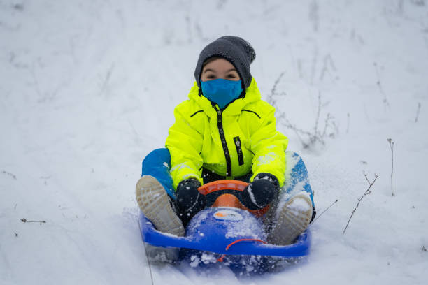 cheerful smiling boy with face mask enjoying time outdoors in snow with bobsled