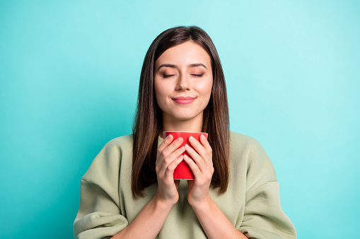 Photo portrait of woman inhaling aroma of nice coffee holding red cup in two hands with closed eyes isolated on vivid turquoise colored background.