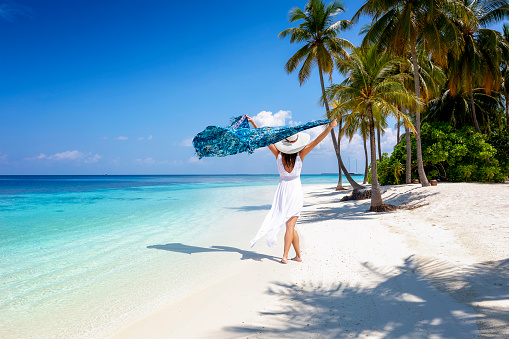 A woman in white summer dress walks on a tropical paradise beach with palm trees and turquoise sea waving a scarf in her hands