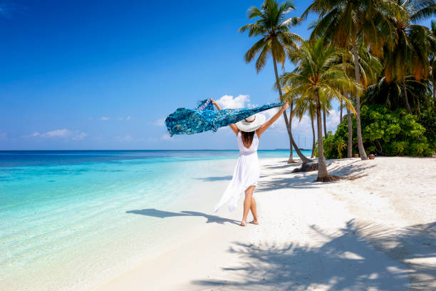 A woman in white summer dress walks on a tropical paradise beach A woman in white summer dress walks on a tropical paradise beach with palm trees and turquoise sea waving a scarf in her hands caribbean islands stock pictures, royalty-free photos & images