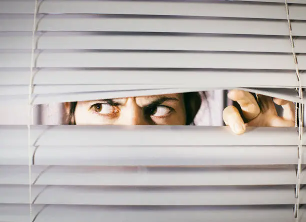 Photo of Young woman peering anxiously through blinds