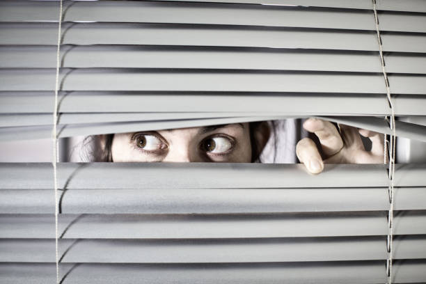 istock Young woman peering anxiously through blinds 1297579706