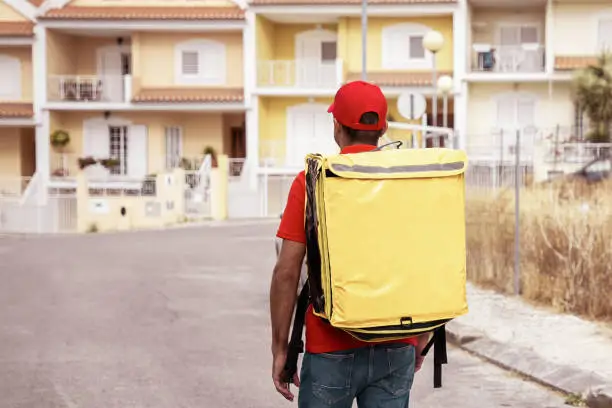 Back view of deliveryman carrying yellow thermo bag. Professional courier walking on street and delivering order on foot. Houses on background. Food delivery service and online shopping concept