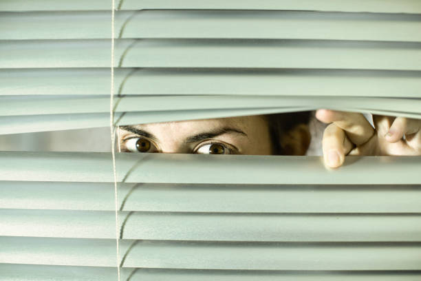 istock Young woman peering anxiously through blinds 1297580019