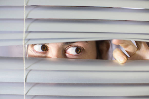 istock Young woman peering anxiously through blinds 1297580835