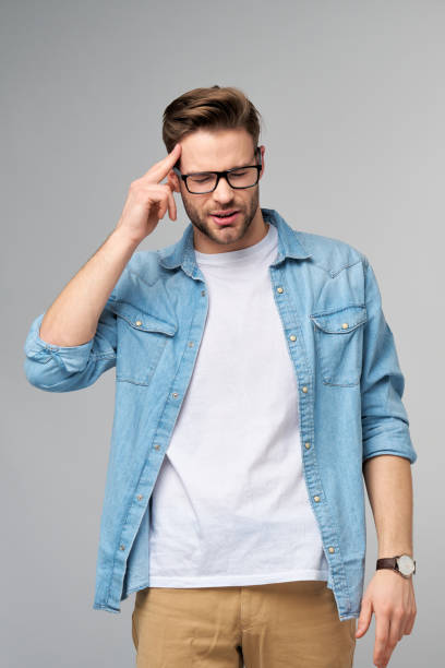 istock Frustrated young caucasian man in jeans shirt touching head with hand while standing overg studio grey background 1297581454
