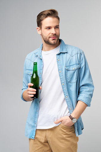 Young Man holding wearing jeans shirt Bottle of beer standing over Grey Background.