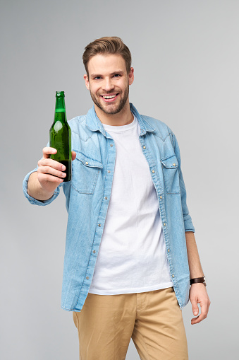 Young Man holding wearing jeans shirt Bottle of beer standing over Grey Background.