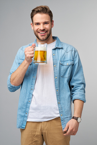 Young Man holding wearing jeans shirt holding glass of beer standing over Grey Background.