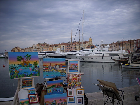 Saint Tropez, France - Mai 05th 2018: The harbor with its expensive yachts and some paintings of a street artist in the foreground at a cloudy day in spring.